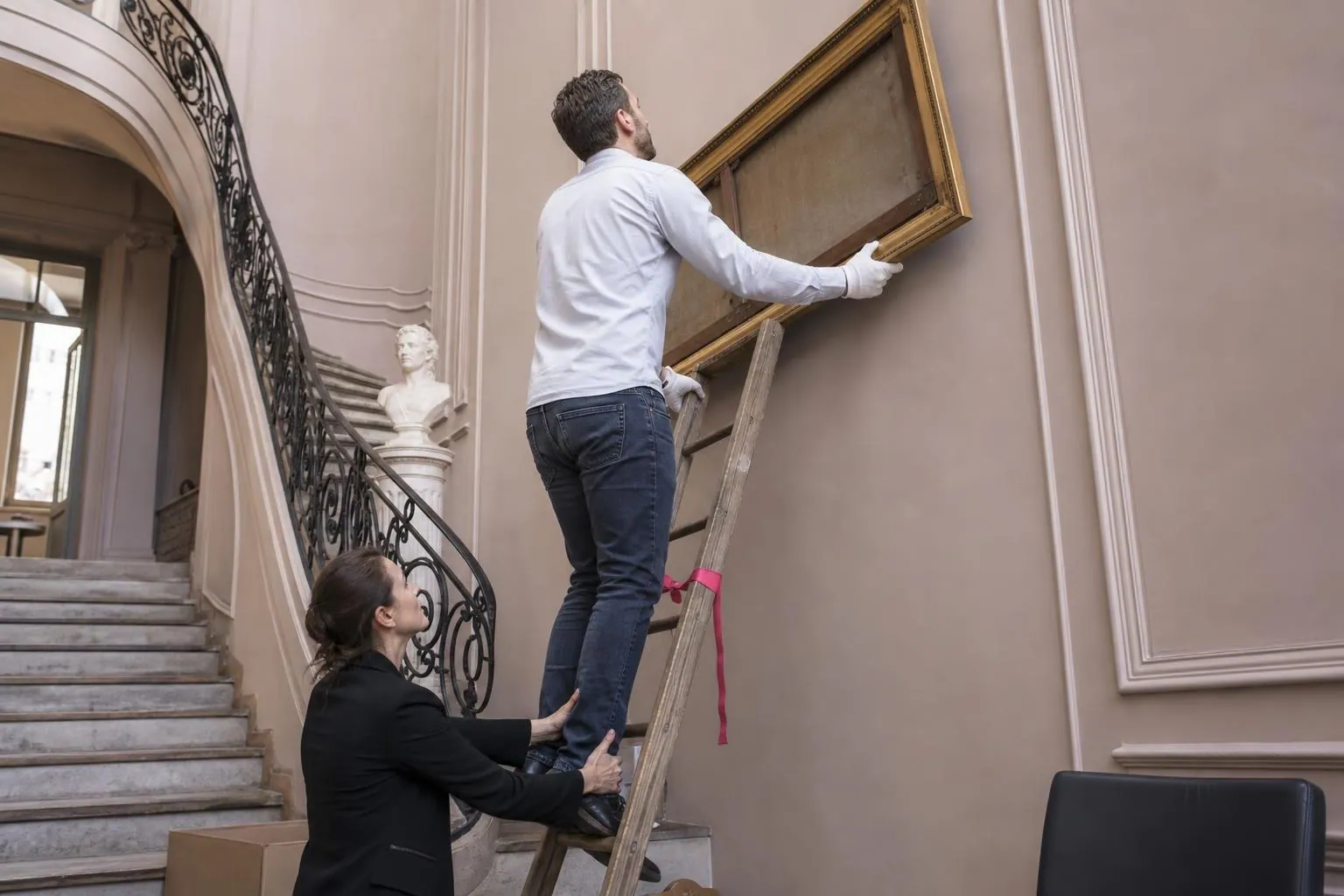 Un agent municipal vérifie sur une échelle un tableau accroché haut dans l’escalier d’honneur d’une mairie