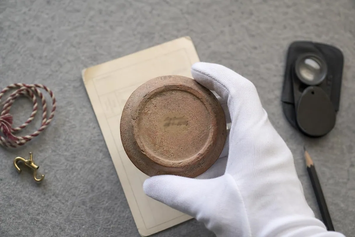 Gros plan d’un numéro d’inventaire manuscrit à l’encre au revers d’un objet de collection posé sur une fiche cartonnée à côté d’une cordelette d’accrochage torsadée